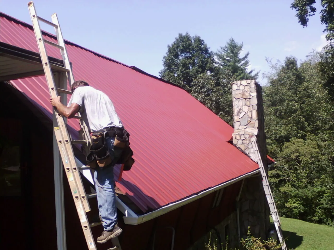 Roofer on a ladder beside a red metal roof and stone chimney — Soffit & Fascia Repair by Roofing Brothers in Ossining